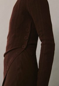 Haut en maille côtelée brun foncé avec une coupe ajustée, comportant des boutons arrondis sur le côté et un tissu légèrement texturé.