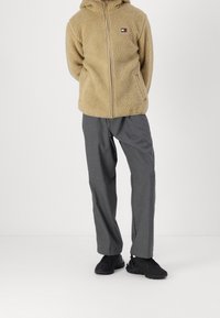 Béžová fleecová mikina s kapucí a zipem vpředu, doplněná o šedé kalhoty a černé sportovní boty. Výrazná textura a ležérní design.