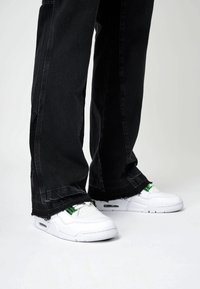 Sneaker bianche con pannelli in mesh testurizzati, accenti verdi e ammortizzazione visibile. Indossate con jeans neri a gamba larga con orli sfrangiati.