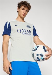 Light gray sports jersey with blue accents, featuring "Qatar Airways" logo, held by a person with a patterned soccer ball in a blue shorts.