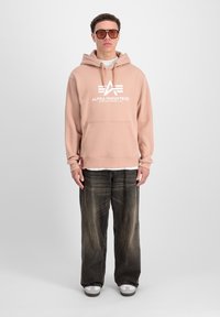 Šviesiai rožinė kapšas su baltu "Alpha Industries" logotipu, priekinė kišenė, uždėta ant balto marškinėlio, derinama su tamsiomis, šiek tiek nusidėvėjusiomis džinsomis ir baltais sportbačiais.
