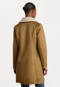 Cappotto marrone con colletto in pelliccia sintetica beige, design dritto, tessuto strutturato e cuciture visibili lungo le giunture. Presenta maniche lunghe e una lunghezza intermedia.