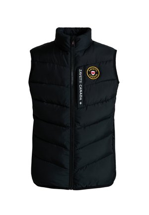Kind met gevlochten haar, draagt een zwart gewatteerd vest, een zwarte trui, een zwarte broek en zwarte sneakers, staand tegen een grijze achtergrond.