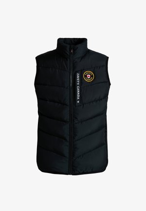 Kind met gevlochten haar, draagt een zwart gewatteerd vest, een zwarte trui, een zwarte broek en zwarte sneakers, staand tegen een grijze achtergrond.