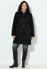 Cappotto di lana nera con collare, chiusura a bottoni e due tasche frontali. Indossato sopra jeans grigi con dettagli borchiati e stivali neri fino alla caviglia.