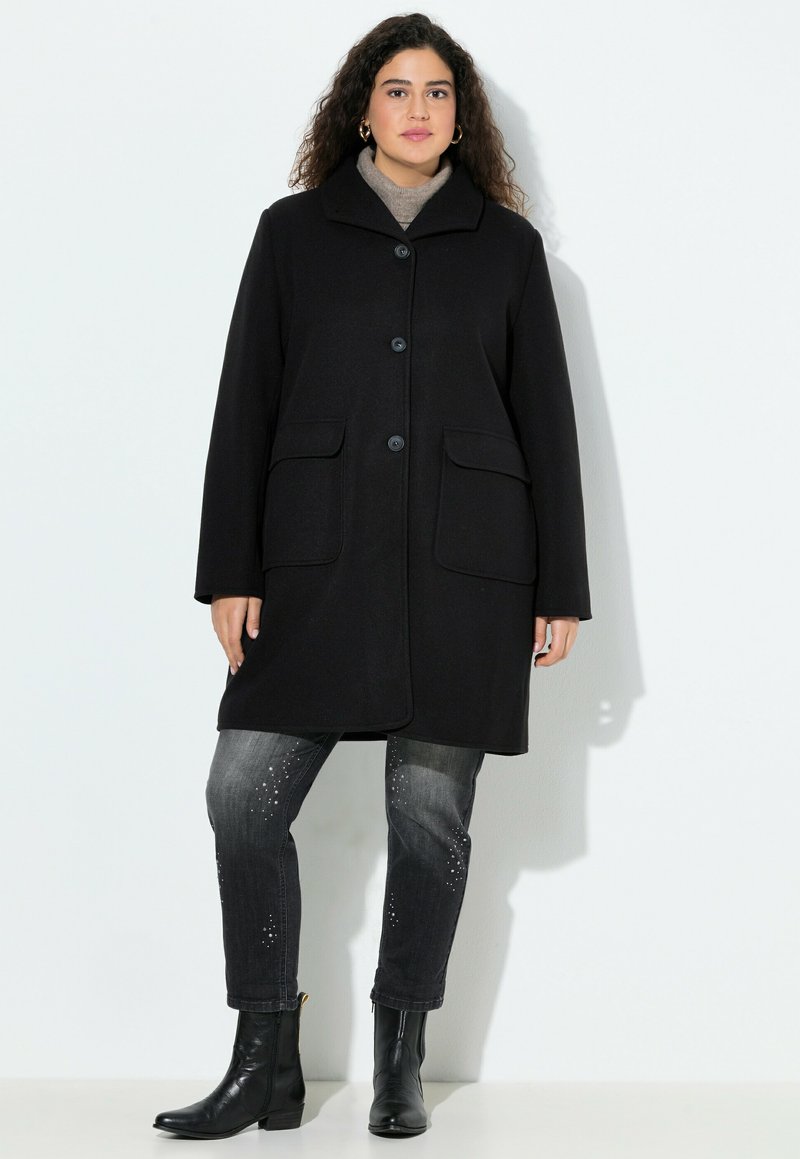 Cappotto di lana nera con collare, chiusura a bottoni e due tasche frontali. Indossato sopra jeans grigi con dettagli borchiati e stivali neri fino alla caviglia.