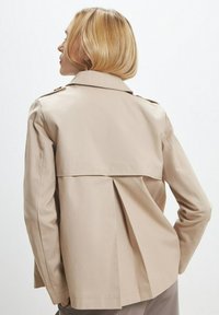 Giacca trench beige con design a doppio strato nella parte posteriore, dettagli con bottoni sulle spalle e texture liscia. Vestibilità sartoriale con linee pulite.