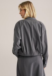 Grå kort jacka med en avslappnad passform, som har en krage, elastisk midja och långa ärmar med knappramar.