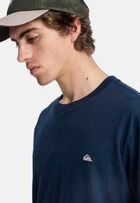 Mørkeblå t-shirt med rund halsudskæring, korte ærmer og struktureret stof, med et lille hvidt logo på brystet.