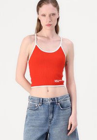 Crop top rouge à côtes avec bord blanc, doté de fines bretelles et d'un logo. Associé à un jean taille haute délavé.