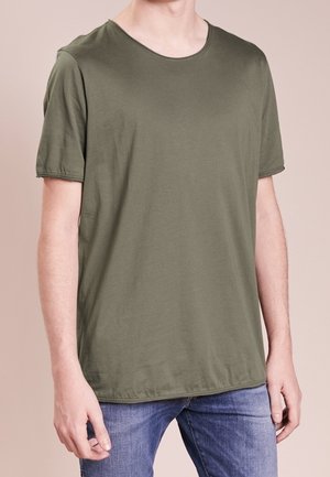 Man wearing plain olive green short-sleeve t-shirt and blue jeans, standing against a light beige background.