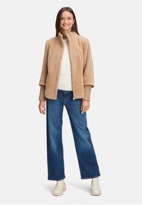 Kobieta uśmiechająca się, ubrana w beżowy, futrzany żakiet, biały sweter, niebieskie szerokie dżinsy i kremowe botki za kostkę, stojąca na białym tle.
