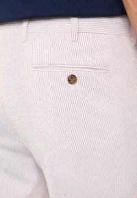 Pantaloni bianchi in cotone con righe rosse verticali, dotati di una tasca posteriore fissata con un bottone marrone. Tessuto a trama, design su misura.
