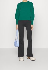 Pull vert en tricot avec manches bouffantes, pantalon noir évasé, sac à main bleu clair et chaussures compensées beige.