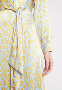 Vaaleansininen mekko, jossa on keltainen leopardikuvio, joka on sidottu vyötärölle pitkällä nauhalla. Pehmeä kangas, jossa on sileä rakenne ja pitkät hihat.