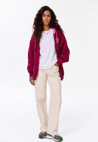 Vínový zapínací svetr přes bílý tričko, béžové široké kalhoty a boty s zeleno-černým vzorem. Kadeřavé vlasy, postoj vestoje.