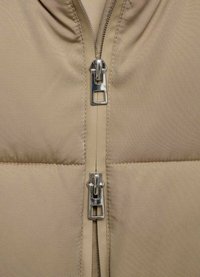 Beige pufferjack close-up met een dubbele rits, metalen hardware en een gladde stof, met de nadruk op de gestikte textuur.