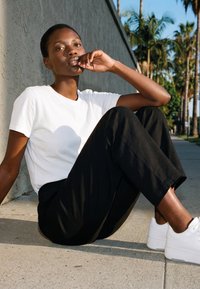 Junge Person sitzt auf dem Bürgersteig an einer Wand, trägt ein weißes Hemd, schwarze Hose und weiße Turnschuhe, mit Palmen im Hintergrund.