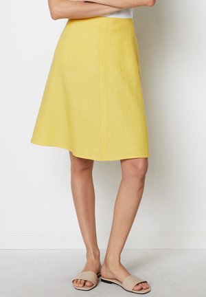 Femme portant une jupe jaune jusqu'aux genoux et des sandales beige à enfiler, debout les bras croisés contre un mur blanc.