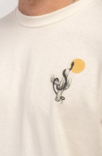 T-shirt en coton beige clair avec une illustration noire d'un cactus enroulé autour d'un serpent et d'un soleil jaune. Encolure ronde, manches courtes.