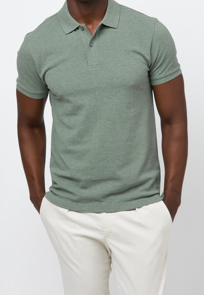 Homme portant un polo vert sauge ajusté à manches courtes et un pantalon écru, debout les mains dans les poches sur fond blanc.