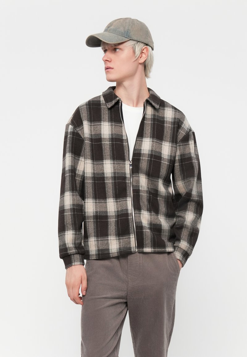 Giacca a zip in plaid nero e beige con colletto, realizzata in tessuto testurizzato. Abbinata a pantaloni in cotone corduroy grigi chiari e un cappellino grigio.