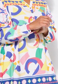 Tunique multicolore présentant un col avec boutons, des motifs géométriques abstraits et des poignets texturés, avec un ourlet inférieur à motifs en bleu.