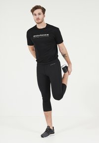 Svart atletisk t-shirt med "ENDURANCE" grafik, tillsammans med svarta capri leggings. Modellen sträcker ut sig, iklädd grå löparskor.