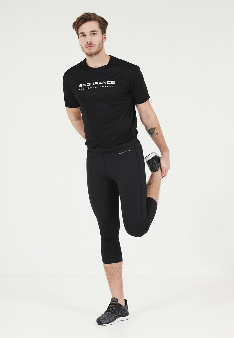 Svart atletisk t-shirt med "ENDURANCE" grafik, tillsammans med svarta capri leggings. Modellen sträcker ut sig, iklädd grå löparskor.