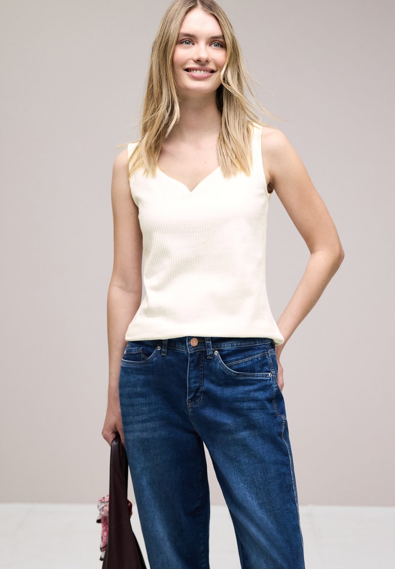 Femme souriante aux cheveux blonds porte un haut blanc sans manches et un jean bleu, tenant un sac de couleur foncée avec une écharpe rose.