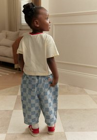 Enfant portant une chemise à manches courtes de couleur crème avec des bretelles rouges et un pantalon large à carreaux bleus, associé à des chaussures rouges et des chaussettes blanches.