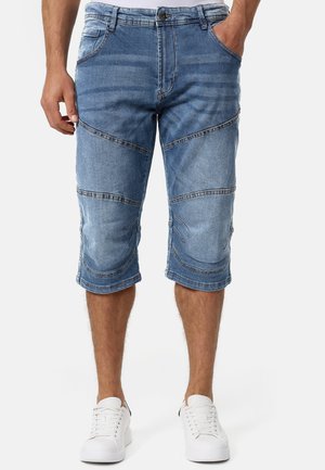 Mand iført blå cowboyshorts til knæet med synlige paneldetaljer og hvide sneakers, stående med hænderne i lommerne.