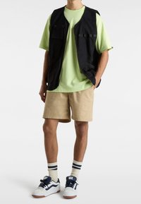 Chaleco negro con cremallera y dos bolsillos delanteros sobre una camiseta verde claro, pantalones cortos beige y zapatillas blancas con detalles azules y calcetines a rayas.