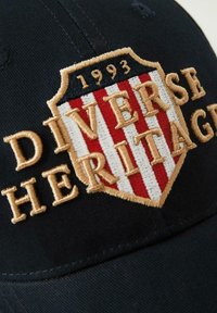 Czarna czapka z haftowanym wzorem w kształcie tarczy, z czerwono-białymi pasami, złotymi literami oraz napisem "1993" nad słowami "Różnorodne Dziedzictwo."