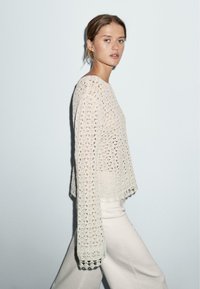 Helles beige Strickpullover mit einem lockeren, offenen Muster und langen Ärmeln, mit gewellten Kanten am Saum und an den Bündchen.