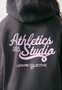 Mørk grå hættetrøje med pink skrift, der siger "Athletics Studio," "Santa Monica," og "Leisure Collective 87" i forskellige skrifttyper.