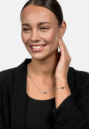Silver jewelry set featuring a heart-shaped pendant necklace and matching bracelet. Chain links and smooth texture. Model wears a black outfit.