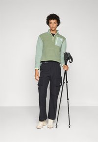 Lichtgroene fleece pullover met halve rits, borstzak; gecombineerd met zwarte cargobroeken, beige wandelschoenen en trekkingstokken.