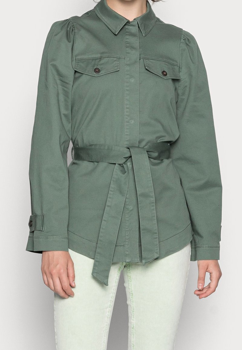 Veste verte ceinturée à manches longues avec poches poitrine boutonnées, portée sur un pantalon clair sur une personne cadrée au niveau du cou.