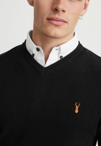 Sort tekstureret sweater med V-udskæring, båret over en hvid skjorte med krave; har et lille orange hjortelogo på den venstre front.