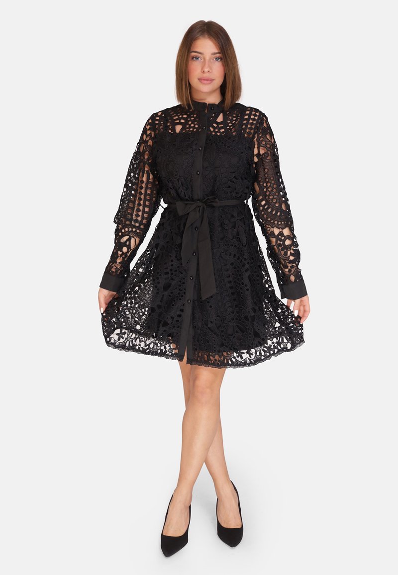 Abito in pizzo nero con maniche lunghe, davanti abbottonato e una cintura in vita. Presenta motivi intricati e una gonna svasata.
