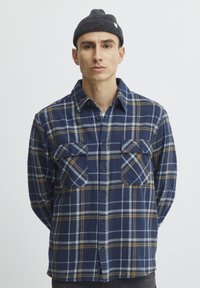 Camisa de franela a cuadros en tonos azul marino con acentos en amarillo y azul claro, cierre de botones en la parte delantera, dos bolsillos en el pecho y un tejido suave y texturizado.
