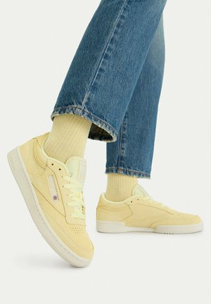 Persona che indossa scarpe da ginnastica Reebok gialle con calzini gialli abbinati e jeans blu su uno sfondo chiaro.