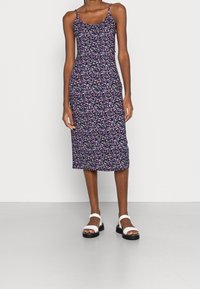 Nainen, jolla on yllään kukkakuosiset, olkaimettomat midi-mekot, joissa on ohuet hihat, sekä valkoiset avokärkiset sandaalit, joissa on paksut mustat pohjat.