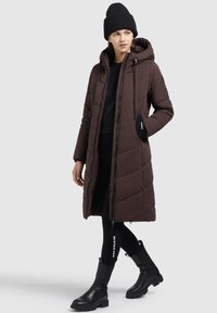 Bruine, gewatteerde lange jas met een capuchon en een rits aan de voorkant, gedragen over een zwarte trui. Een zwarte geribde beanie en enkellaarzen maken de look compleet.