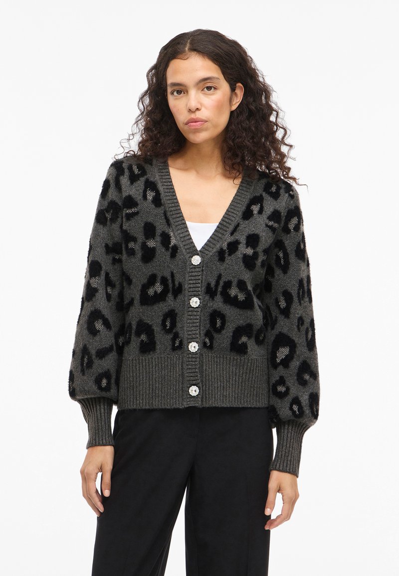 Γκρι καμπαρντίνα με μαύρο λεοπάρ σχέδιο, υφή υφάσματος, V-λαιμό και κλείσιμο με έξι κουμπιά. Οι ριγωτές μανσέτες και το στρίφωμα προσθέτουν αντίθεση.