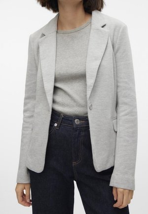 Kvinde iført en lys grå blazer over en grå rundhalset top, kombineret med højtaljede mørkeblå jeans mod en ensfarvet baggrund.