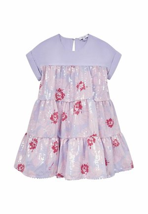 Ung pige iført en lavendel- og lyserød lagdelt kjole med blomsterprint og gyldne sandaler, stående mod en ensfarvet lys baggrund.