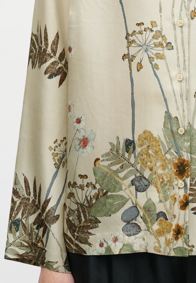 Blouse à motif floral avec un fond beige, présentant des feuilles vertes et des fleurs multicolores. Confectionnée en tissu lisse et léger avec une fermeture boutonnée à l'avant.