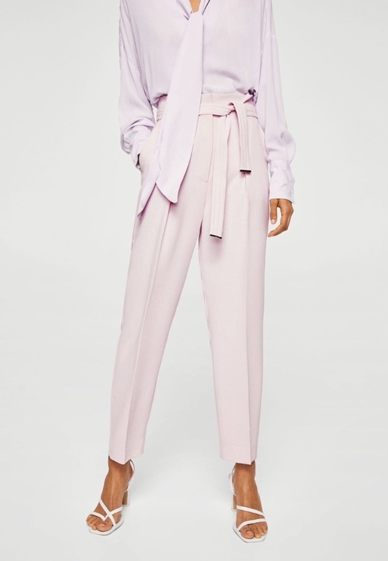 Lichtroze op maat gemaakte broek met een hoge taille, riemdetail en een taps toelopende fit. Gecombineerd met een lavendelkleurige blouse met lange mouwen en witte sandalen.
